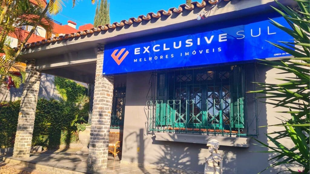 Fachada da imobiliária Exclusive Sul Melhores Imóveis em um dia ensolarado. A casa térrea possui pintura cinza, janelas com venezianas verdes, varanda sustentada por pilares de pedra e telhado de cerâmica. O destaque é a placa azul com letras brancas com o logotipo e nome da empresa.