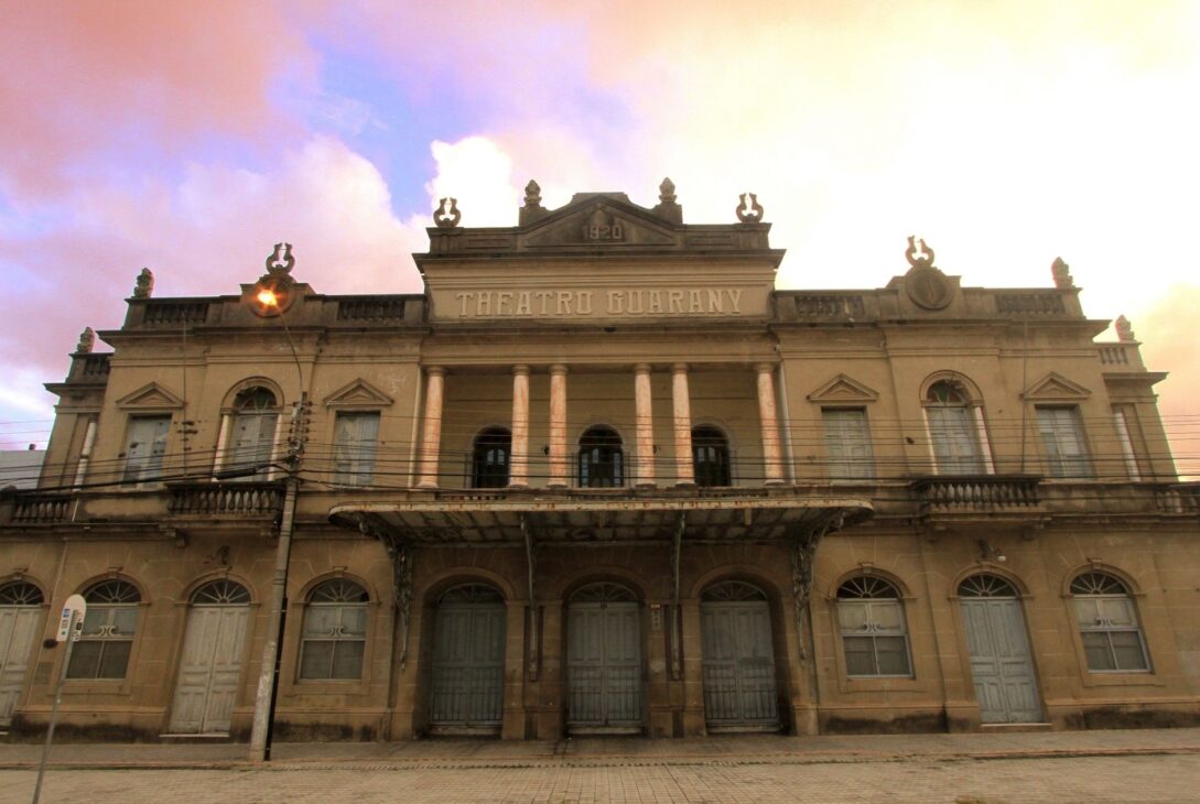 Fachada histórica do Theatro Guarany em Pelotas sob um céu de fim de tarde, destacando a arquitetura neoclássica e colunas frontais.