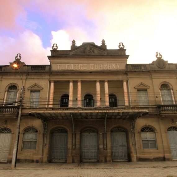 Fachada histórica do Theatro Guarany em Pelotas sob um céu de fim de tarde, destacando a arquitetura neoclássica e colunas frontais.