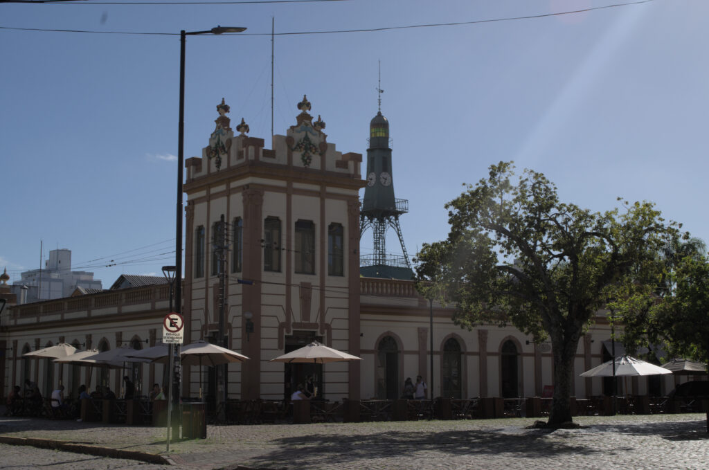 Prédio histórico do Mercado Central de Pelotas com mesas externas sob ombrelones e a icônica torre do relógio ao fundo.