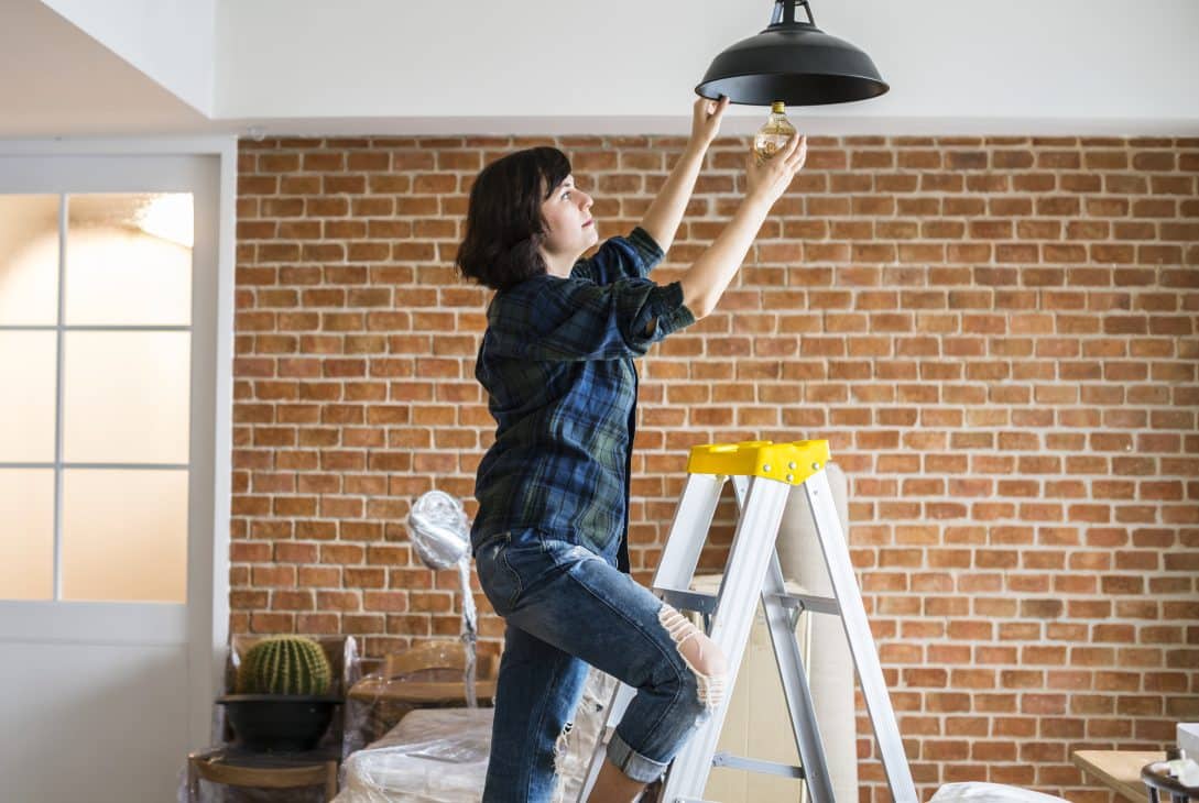 Mulher subindo em uma escada para trocar uma lâmpada em uma luminária suspensa, em um ambiente interno com parede de tijolos aparentes. O cenário sugere uma atividade de manutenção ou melhorias em uma casa ou apartamento.