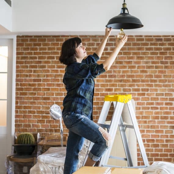 Mulher subindo em uma escada para trocar uma lâmpada em uma luminária suspensa, em um ambiente interno com parede de tijolos aparentes. O cenário sugere uma atividade de manutenção ou melhorias em uma casa ou apartamento.