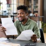 documentacao-necessaria-para-comprar-um-imovel-homem-jovem-negro-sorrindo-olhando-para-os-documentos-que-esta-segurando