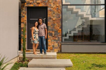 Casal em frente à fachada de uma casa moderna com revestimento em pedra natural e grande vitrine de vidro, simbolizando a conquista da casa própria em Pelotas.
