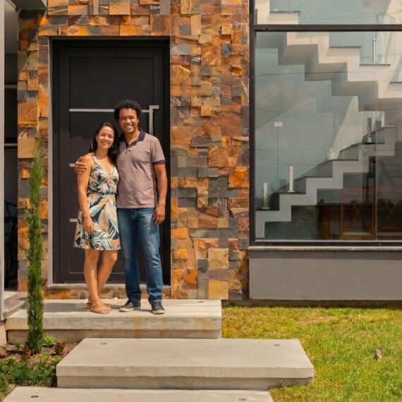 Casal em frente à fachada de uma casa moderna com revestimento em pedra natural e grande vitrine de vidro, simbolizando a conquista da casa própria em Pelotas.