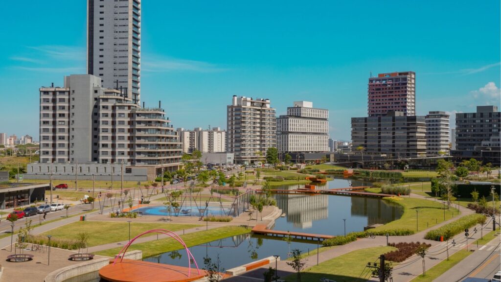 Vista panorâmica do Parque Una em Pelotas, destacando o lago central, passarelas de madeira, áreas verdes e edifícios modernos de alto padrão ao fundo sob céu azul.