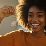 A imagem retrata uma jovem sorridente segurando um chaveiro com a miniatura de uma casa e duas chaves, simbolizando a conquista da casa própria. Ela veste uma blusa de manga comprida em tom mostarda e transmite alegria e realização, com a luz do sol iluminando seu rosto e criando um clima acolhedor e esperançoso. O fundo desfocado sugere um ambiente interno bem iluminado, reforçando a ideia de um novo lar. A cena representa um momento marcante e positivo, frequentemente associado à compra de um imóvel, possivelmente com apoio de financiamento ou uso do FGTS.