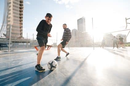 Dois jogadores disputando uma partida de futebol em uma quadra ao ar livre, com outras pessoas ao fundo. A cena acontece em um ambiente urbano, com prédios modernos e estruturas em construção ao fundo, iluminados por uma luz do sol intensa.