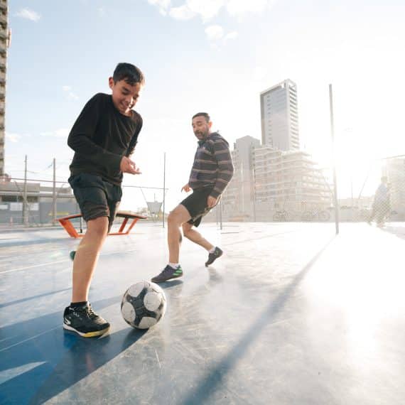 Dois jogadores disputando uma partida de futebol em uma quadra ao ar livre, com outras pessoas ao fundo. A cena acontece em um ambiente urbano, com prédios modernos e estruturas em construção ao fundo, iluminados por uma luz do sol intensa.