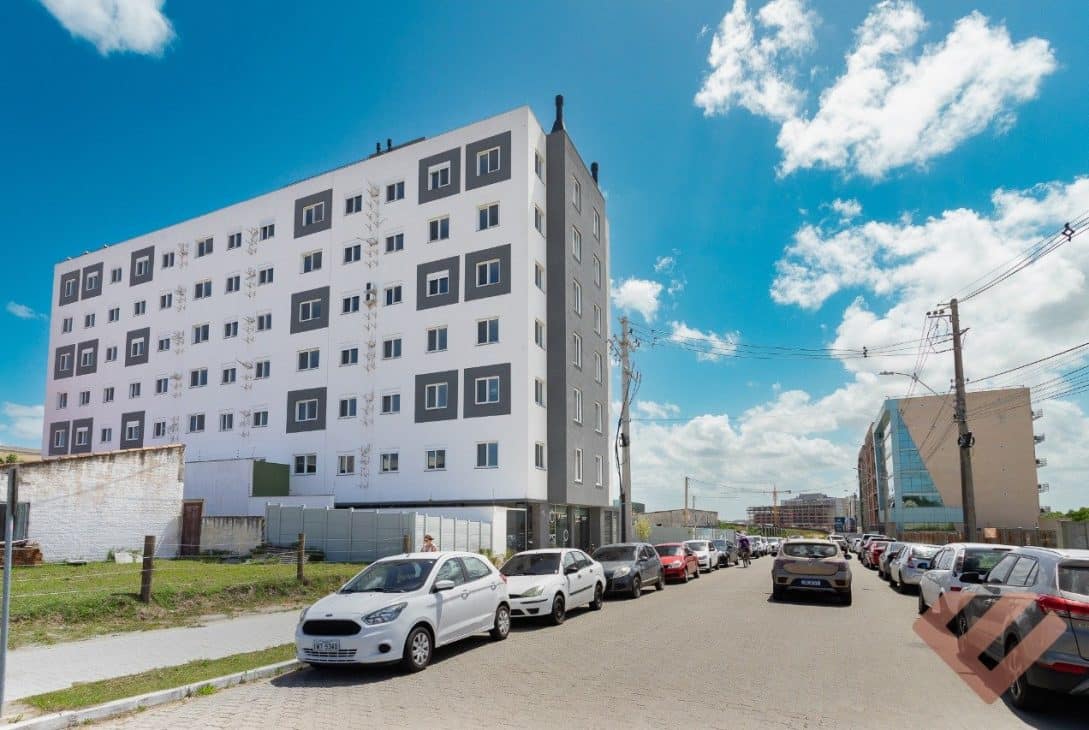 Imagem de um edifício residencial de vários andares, com fachada branca e detalhes em cinza nas janelas. O prédio está localizado em uma rua de calçamento de paralelepípedos, onde carros estão estacionados ao longo dos dois lados. O céu está claro e ensolarado, com algumas nuvens brancas. Ao fundo, vêem-se outras construções e alguns postes de energia com fiação aparente.