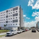Imagem de um edifício residencial de vários andares, com fachada branca e detalhes em cinza nas janelas. O prédio está localizado em uma rua de calçamento de paralelepípedos, onde carros estão estacionados ao longo dos dois lados. O céu está claro e ensolarado, com algumas nuvens brancas. Ao fundo, vêem-se outras construções e alguns postes de energia com fiação aparente.