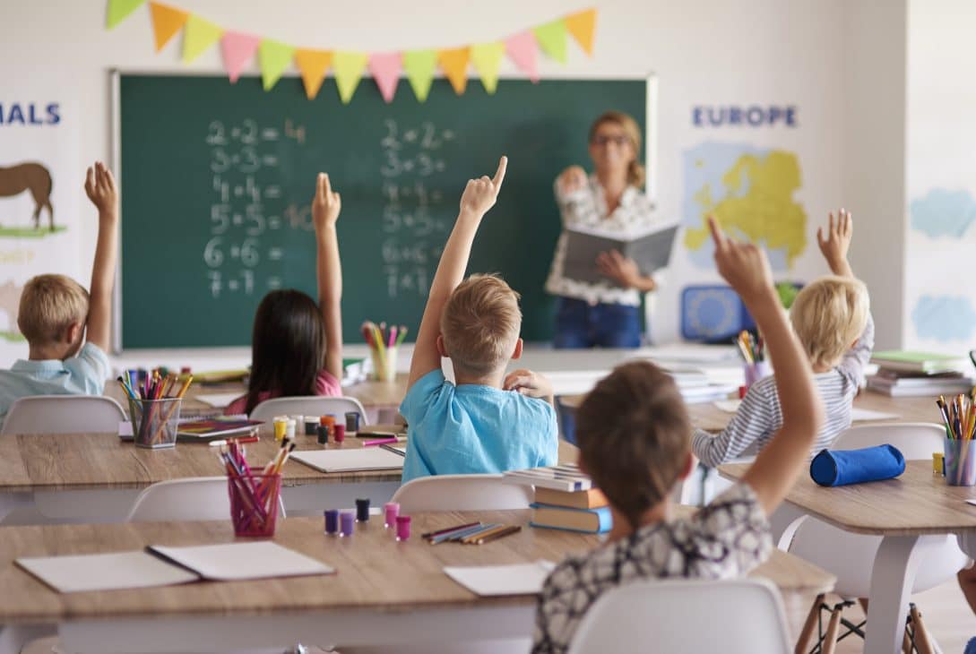 ala de aula com crianças sentadas em carteiras, levantando as mãos para responder a perguntas da professora. Ao fundo, a professora está em pé, próxima a um quadro verde com operações matemáticas escritas.
