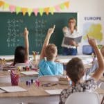 ala de aula com crianças sentadas em carteiras, levantando as mãos para responder a perguntas da professora. Ao fundo, a professora está em pé, próxima a um quadro verde com operações matemáticas escritas.