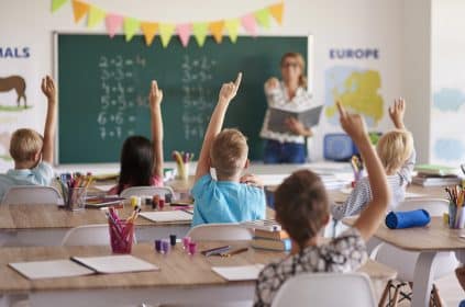 ala de aula com crianças sentadas em carteiras, levantando as mãos para responder a perguntas da professora. Ao fundo, a professora está em pé, próxima a um quadro verde com operações matemáticas escritas.