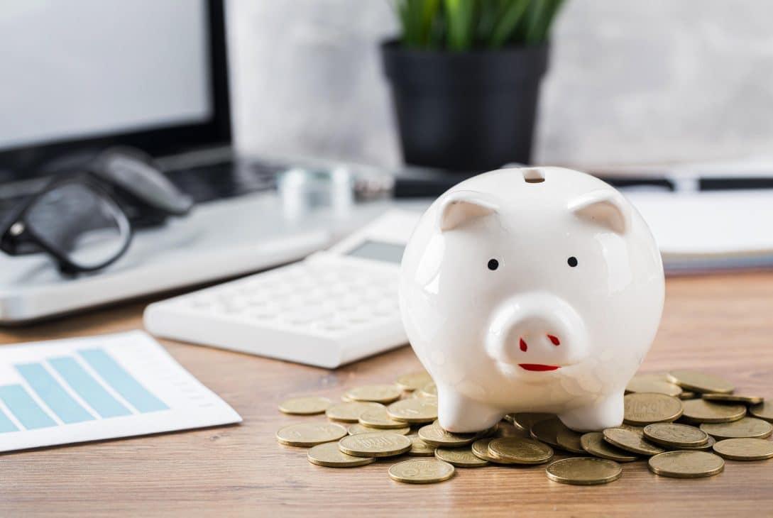 Cofrinho em formato de porquinho branco sobre uma mesa de madeira, cercado por moedas douradas. Ao fundo, é possível ver um laptop, óculos, uma calculadora e gráficos, sugerindo planejamento financeiro e economia.