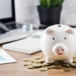 Cofrinho em formato de porquinho branco sobre uma mesa de madeira, cercado por moedas douradas. Ao fundo, é possível ver um laptop, óculos, uma calculadora e gráficos, sugerindo planejamento financeiro e economia.