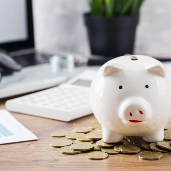 Cofrinho em formato de porquinho branco sobre uma mesa de madeira, cercado por moedas douradas. Ao fundo, é possível ver um laptop, óculos, uma calculadora e gráficos, sugerindo planejamento financeiro e economia.