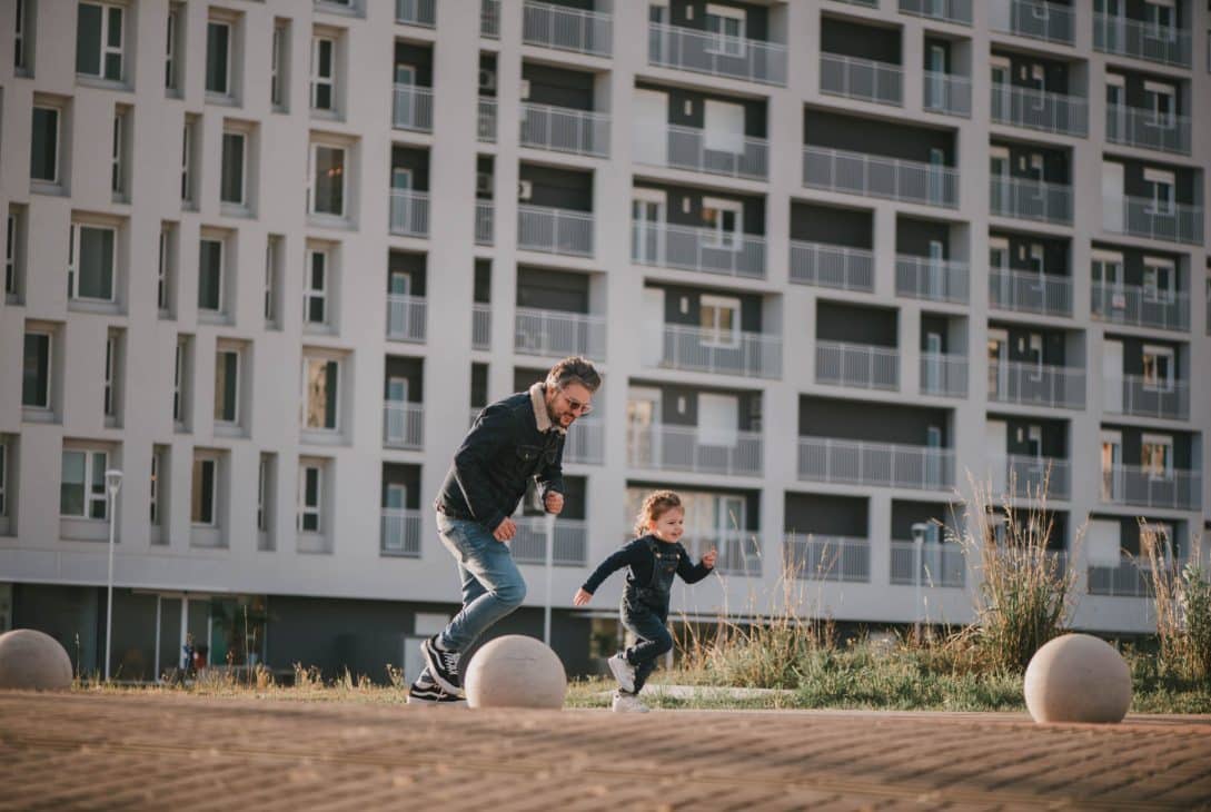 Homem brincando com uma criança em um espaço ao ar livre, com um edifício residencial moderno ao fundo, simbolizando a convivência em comunidades urbanas."