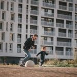 Homem brincando com uma criança em um espaço ao ar livre, com um edifício residencial moderno ao fundo, simbolizando a convivência em comunidades urbanas."
