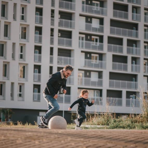 Homem brincando com uma criança em um espaço ao ar livre, com um edifício residencial moderno ao fundo, simbolizando a convivência em comunidades urbanas."