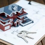 Uma miniatura de casa vermelha com telhado azul está posicionada sobre um projeto arquitetônico impresso em uma prancheta de madeira. Ao lado, um conjunto de chaves prateadas simboliza um contrato de locação ou compra de imóvel.