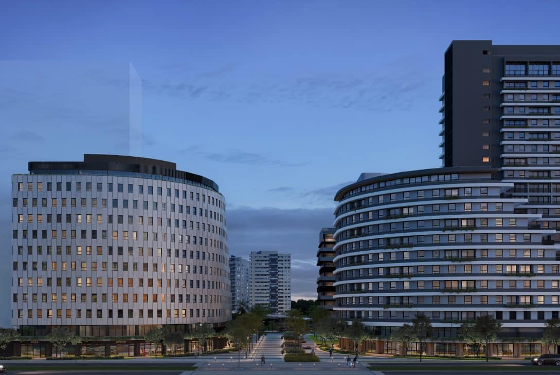 A imagem mostra uma vista da fachada de dois prédios modernos, com arquitetura arrojada, em uma área urbana. Os edifícios têm formas curvas e são compostos por vários andares com janelas grandes, proporcionando uma sensação de elegância e sofisticação. O cenário é ao entardecer, com a iluminação interna visível nas janelas, destacando-se sobre o céu azul suave. Em frente aos prédios, há uma área ajardinada com caminhos, árvores e espaços públicos bem cuidados, contribuindo para a atmosfera agradável e urbanizada. A perspectiva mostra a entrada principal, com espaço para pedestres e veículos.