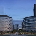 A imagem mostra uma vista da fachada de dois prédios modernos, com arquitetura arrojada, em uma área urbana. Os edifícios têm formas curvas e são compostos por vários andares com janelas grandes, proporcionando uma sensação de elegância e sofisticação. O cenário é ao entardecer, com a iluminação interna visível nas janelas, destacando-se sobre o céu azul suave. Em frente aos prédios, há uma área ajardinada com caminhos, árvores e espaços públicos bem cuidados, contribuindo para a atmosfera agradável e urbanizada. A perspectiva mostra a entrada principal, com espaço para pedestres e veículos.