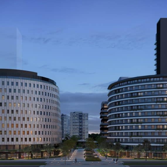 A imagem mostra uma vista da fachada de dois prédios modernos, com arquitetura arrojada, em uma área urbana. Os edifícios têm formas curvas e são compostos por vários andares com janelas grandes, proporcionando uma sensação de elegância e sofisticação. O cenário é ao entardecer, com a iluminação interna visível nas janelas, destacando-se sobre o céu azul suave. Em frente aos prédios, há uma área ajardinada com caminhos, árvores e espaços públicos bem cuidados, contribuindo para a atmosfera agradável e urbanizada. A perspectiva mostra a entrada principal, com espaço para pedestres e veículos.