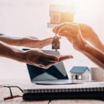 A imagem mostra duas pessoas trocando as chaves de uma casa, simbolizando a negociação ou venda de um imóvel. As mãos de cada pessoa estão estendidas, com a chave de uma casa sendo transferida de uma para a outra. Ao fundo, há um pequeno modelo de casa, um laptop e um caderno, sugerindo que o ambiente é de trabalho, possivelmente um escritório imobiliário.