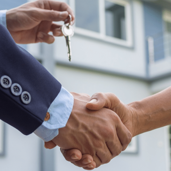 Imagem de duas pessoas apertando as mãos, uma delas está passando uma chave para a outra. Ao fundo, é possível ver uma casa, simbolizando a venda de um imóvel