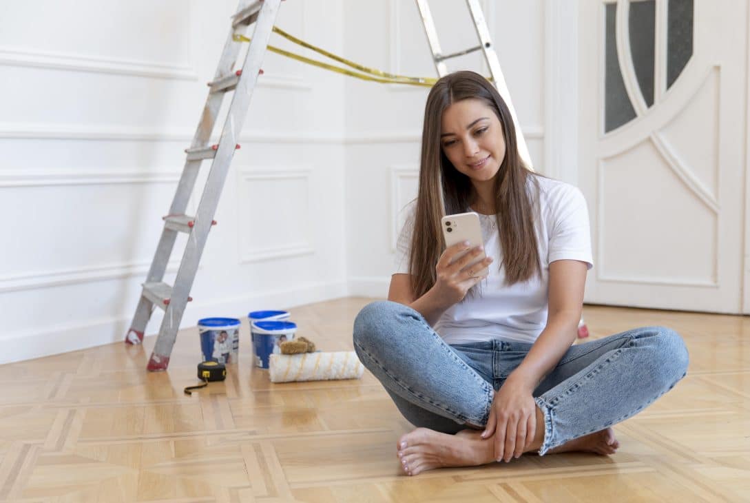 A imagem mostra uma mulher sentada no chão de um ambiente que está passando por uma reforma. Ela está usando uma camiseta branca e jeans claros, sorrindo enquanto olha para seu celular. Ao fundo, há uma escada de alumínio e materiais de pintura, como latas de tinta e um rolo de pintura, sugerindo que ela está em um momento de pausa no trabalho.