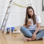 A imagem mostra uma mulher sentada no chão de um ambiente que está passando por uma reforma. Ela está usando uma camiseta branca e jeans claros, sorrindo enquanto olha para seu celular. Ao fundo, há uma escada de alumínio e materiais de pintura, como latas de tinta e um rolo de pintura, sugerindo que ela está em um momento de pausa no trabalho.