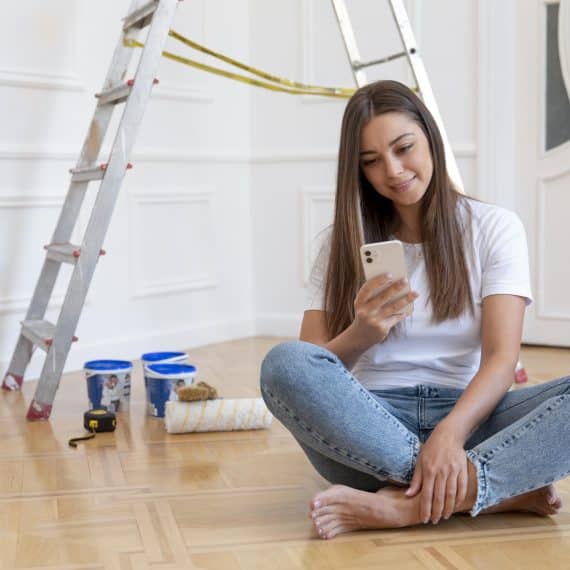 A imagem mostra uma mulher sentada no chão de um ambiente que está passando por uma reforma. Ela está usando uma camiseta branca e jeans claros, sorrindo enquanto olha para seu celular. Ao fundo, há uma escada de alumínio e materiais de pintura, como latas de tinta e um rolo de pintura, sugerindo que ela está em um momento de pausa no trabalho.