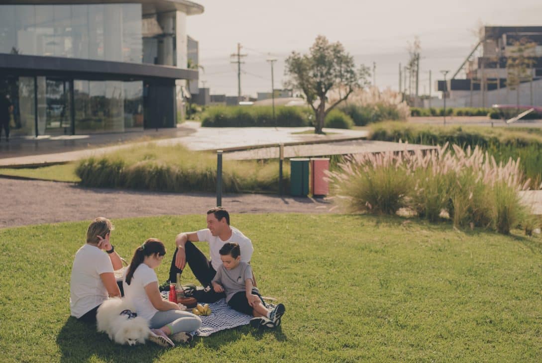 A imagem mostra uma família desfrutando de um momento ao ar livre em um espaço verde, localizado em um ambiente urbano moderno. Eles estão sentados em uma manta sobre a grama, com um cachorro de pelagem branca ao lado. A família consiste em um homem, uma mulher e duas crianças, com todos vestidos de forma casual. Ao fundo, é possível ver uma construção moderna e arquitetonicamente interessante, com árvores e vegetação ao redor.