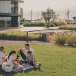 A imagem mostra uma família desfrutando de um momento ao ar livre em um espaço verde, localizado em um ambiente urbano moderno. Eles estão sentados em uma manta sobre a grama, com um cachorro de pelagem branca ao lado. A família consiste em um homem, uma mulher e duas crianças, com todos vestidos de forma casual. Ao fundo, é possível ver uma construção moderna e arquitetonicamente interessante, com árvores e vegetação ao redor.