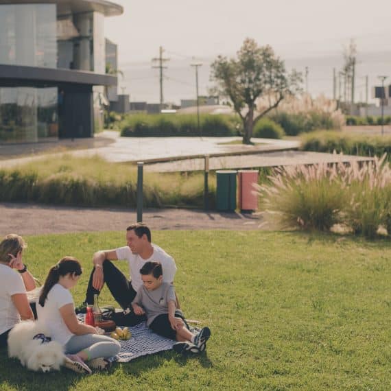 A imagem mostra uma família desfrutando de um momento ao ar livre em um espaço verde, localizado em um ambiente urbano moderno. Eles estão sentados em uma manta sobre a grama, com um cachorro de pelagem branca ao lado. A família consiste em um homem, uma mulher e duas crianças, com todos vestidos de forma casual. Ao fundo, é possível ver uma construção moderna e arquitetonicamente interessante, com árvores e vegetação ao redor.