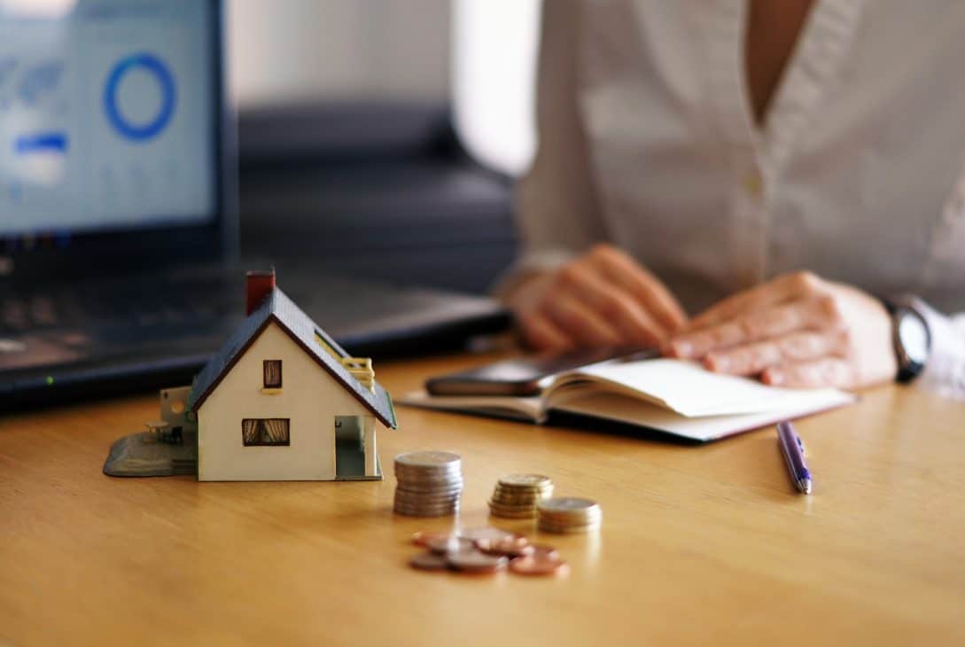 Miniatura de casa ao lado de pilhas de moedas sobre mesa, com pessoa escrevendo em caderno e notebook ao fundo, representando planejamento financeiro imobiliário.
