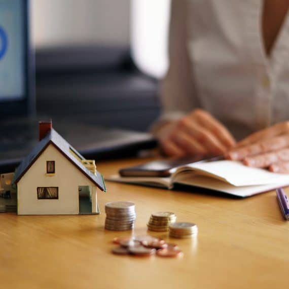 Miniatura de casa ao lado de pilhas de moedas sobre mesa, com pessoa escrevendo em caderno e notebook ao fundo, representando planejamento financeiro imobiliário.