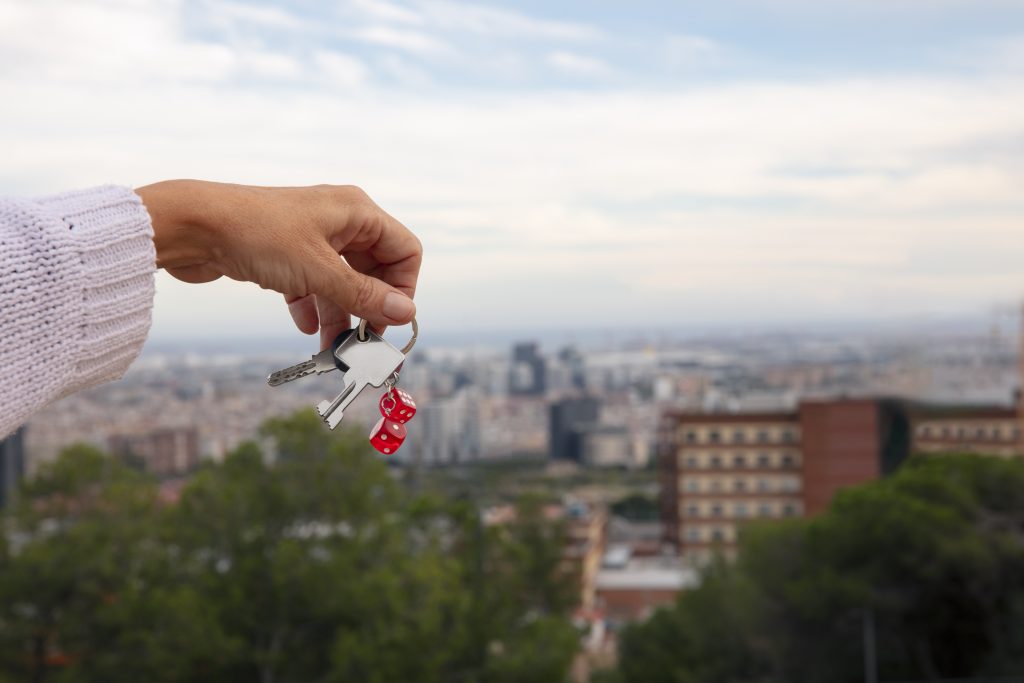 Mão segurando chaves com chaveiro de dados vermelhos, com vista desfocada de cidade ao fundo.