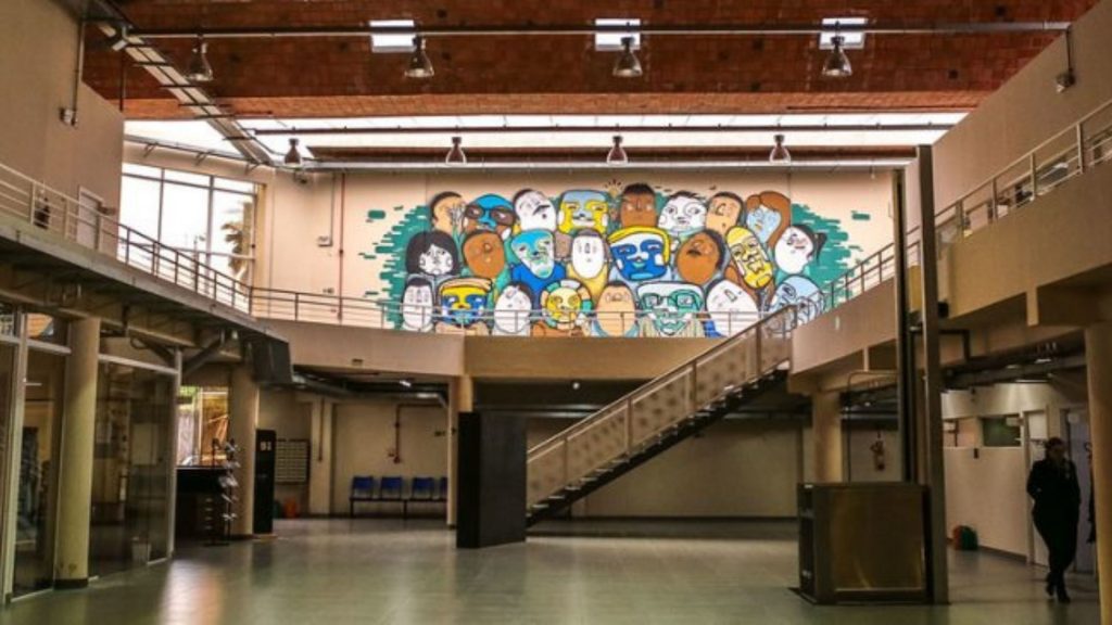 Interior de prédio com pé-direito alto e escadas metálicas, destacando mural colorido com rostos estilizados na parede ao fundo.