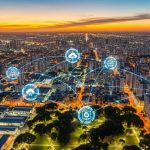 Vista aérea de uma cidade iluminada ao entardecer, com ícones digitais representando tecnologia e conectividade sobrepostos à imagem.