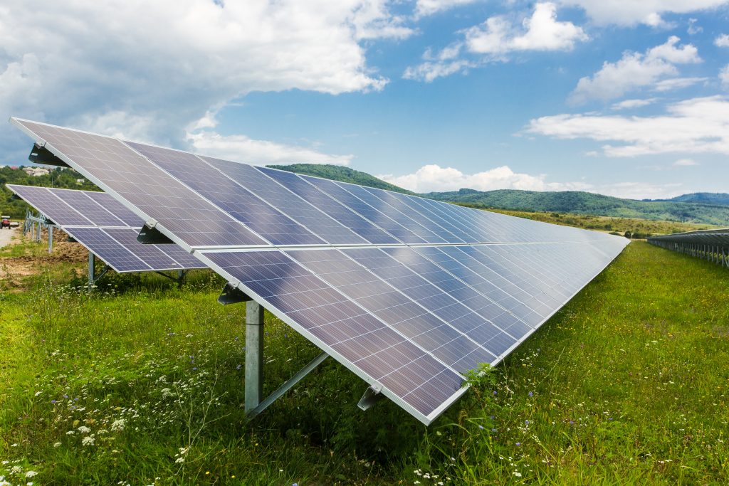 Painéis solares instalados em área verde, com céu parcialmente nublado ao fundo e colinas cobertas por vegetação.