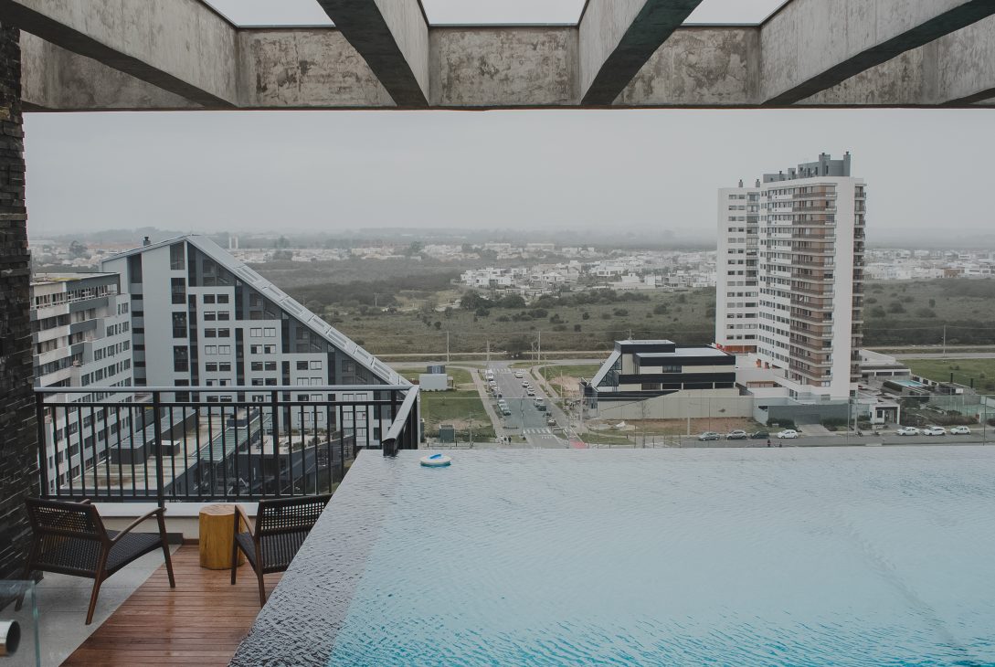 Vista panorâmica de condomínio moderno com piscina de borda infinita na cobertura e prédios residenciais ao fundo.
