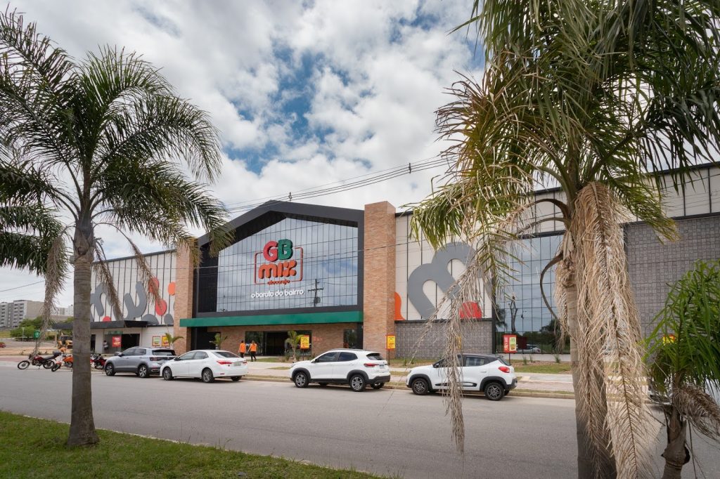 Fachada do GB Mix Atacadista com letreiro colorido e slogan “O barato do bairro”, ladeado por palmeiras e carros estacionados.