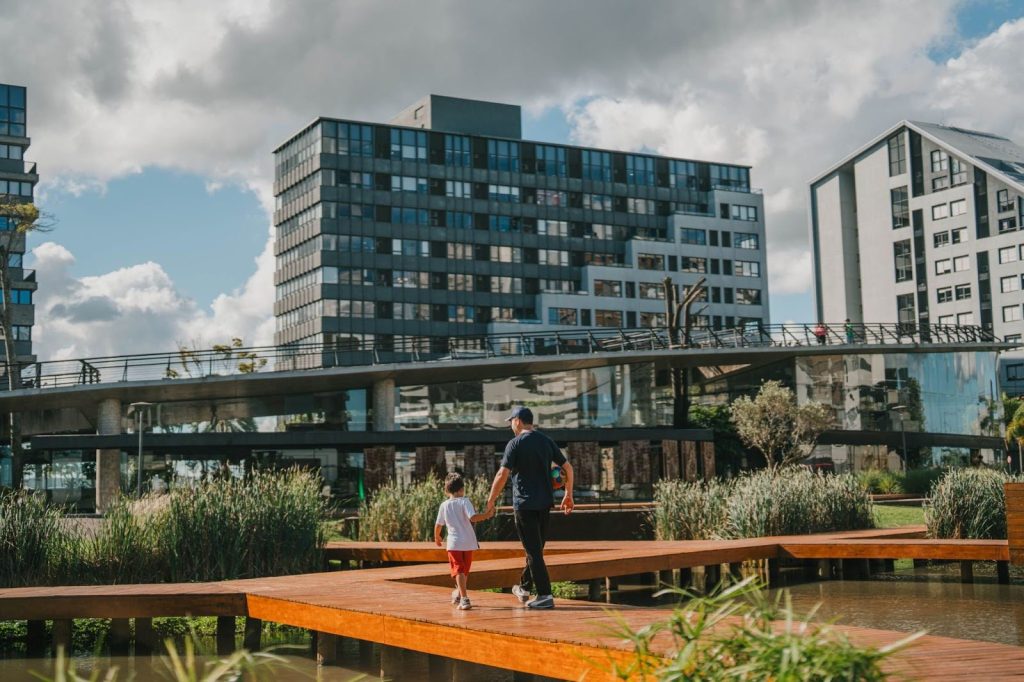 Pai e filho caminhando por passarela de madeira sobre lago em parque urbano, com prédios modernos ao fundo.