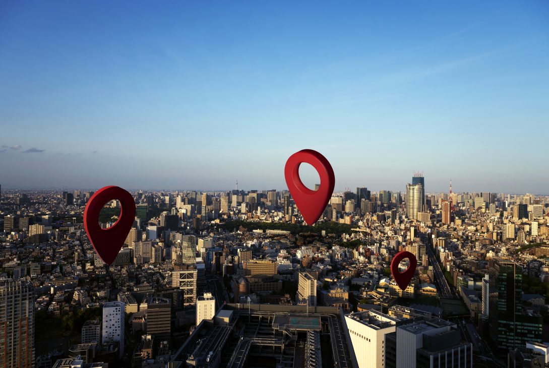 Ilustração com grandes marcadores vermelhos de localização sobrepostos a uma vista aérea de cidade, simbolizando pontos de interesse.