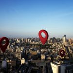 Ilustração com grandes marcadores vermelhos de localização sobrepostos a uma vista aérea de cidade, simbolizando pontos de interesse.