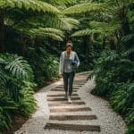 Mulher caminhando por trilha com pedras brancas e tábuas de madeira, cercada por vegetação densa e folhagens grandes, segurando um tapete de yoga enrolado.