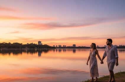 Casal caminhando de mãos dadas à beira de um lago ao entardecer, com o céu colorido em tons de rosa e laranja e a silhueta de prédios ao fundo.