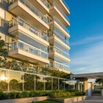 Fachada de prédio residencial de luxo em Pelotas com sacadas de vidro, arquitetura contemporânea e jardim iluminado pelo pôr do sol.